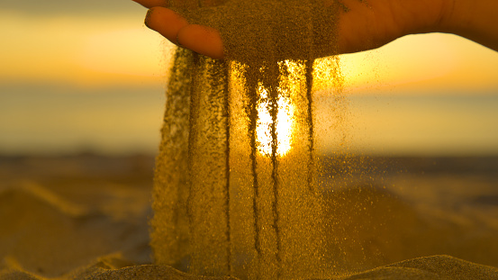 CLOSE UP: Small particles of dry sand fall between gentle unknown female's fingers and back on pretty shore. Beautiful summer sunset illuminates sandy beach and woman sifting dry sand with her hands.