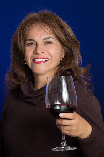 Latin american lady in a studio portrait over blue background. She is in her fifties, has long brown hair, brown eyes. She is smiling and she is offering a toast with a glass of wine.