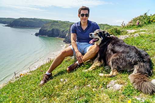 Single Hispanic man lifestyle portrait at beach sitting on headland at Skrinkle Haven on the Pembrokeshire Coast with his dog