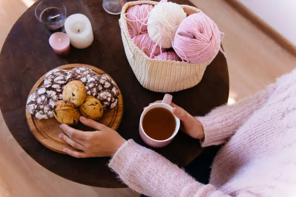 Cup of black tea and plate of fresh homemade cookies on wooden table with woolen coils of thread. Woman hands holding hot aromatic tea. Morning breakfast, warm and cozy