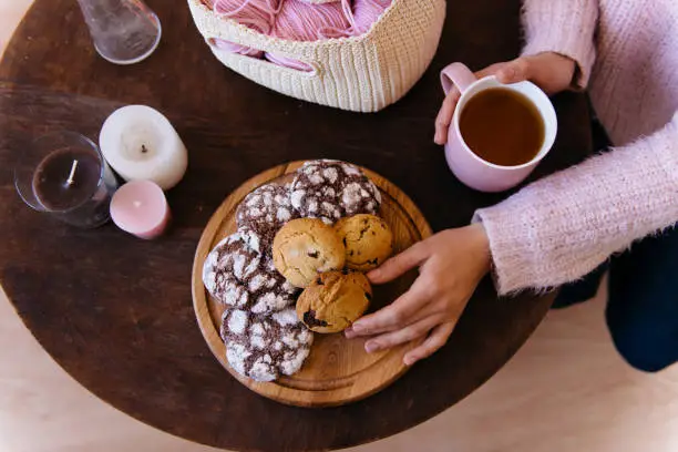 Cup of black tea and plate of fresh homemade cookies on wooden table with woolen coils of thread. Woman hands holding hot aromatic tea. Morning breakfast, warm and cozy