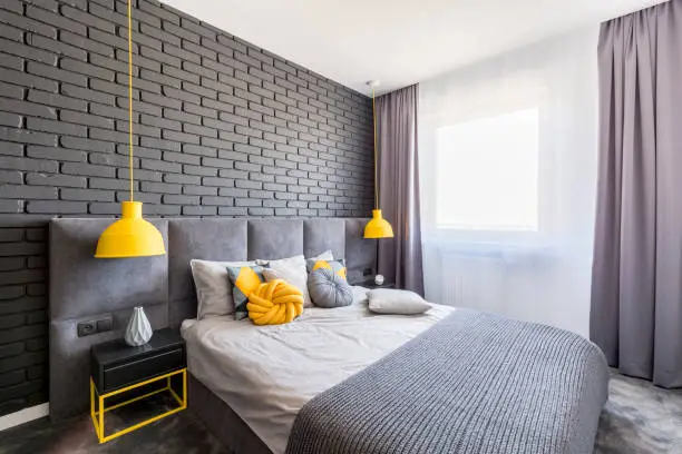 Photo of Real photo of grey and yellow bedroom interior with window with curtains, brick wall, two lamps and knot cushion