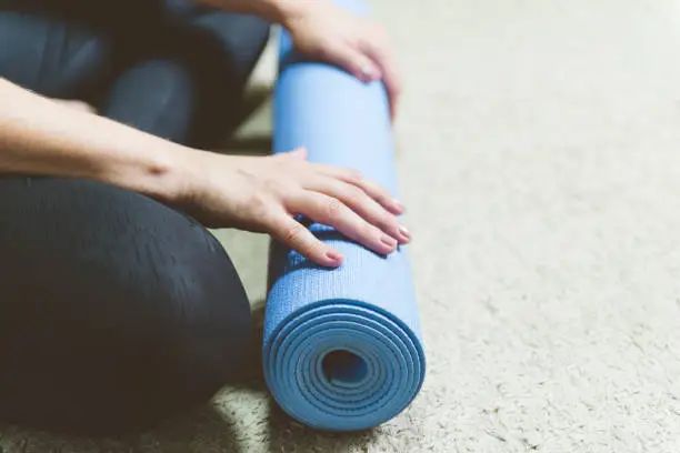 Caucasian woman in black clothes sitting on bege carpet rolling blue yogamat