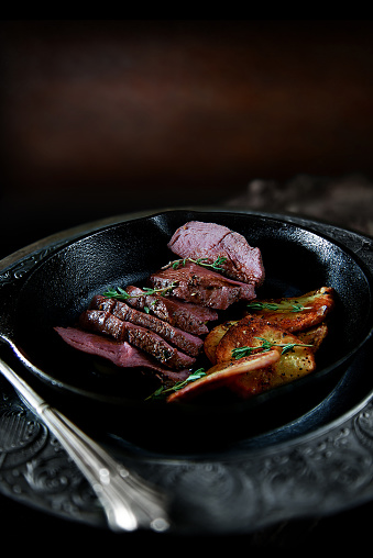 Pan-fried wild venison fillet with fried potato slices and thyme herbs. Shot against a dark, rustic background with generous accommodation for copy space.