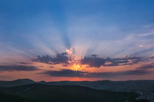 Photo of The dawn sky, the rays of the rising sun Shine through the clouds against the blue sky. At the bottom of the sleeping forest, mountains and the village. Rays of God.
