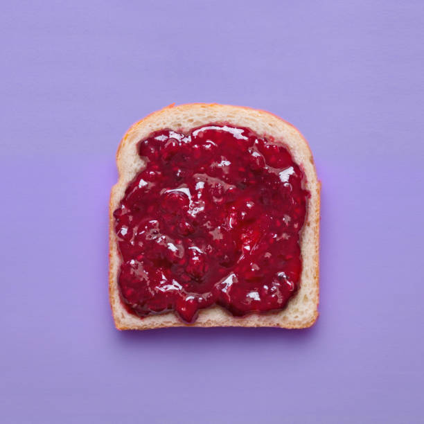 istock Toast slice with fruit jam on a purple background viewed from above. Slice of bread with raspberry marmalade isolated on a colorful background. Top view 1009431430