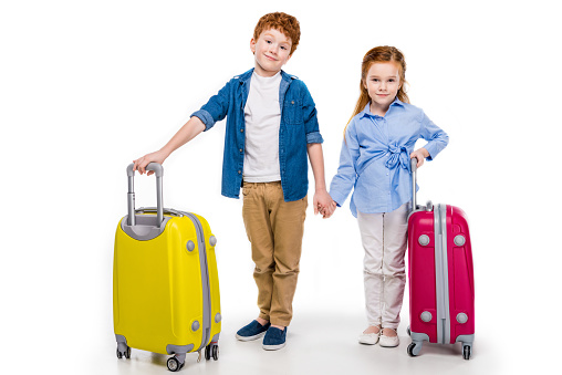 cute little kids holding hands while standing with suitcases and smiling at camera isolated on white