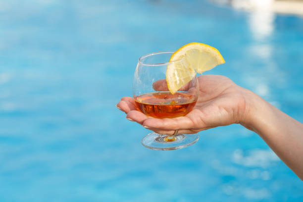 Glass of fragrant cognac with a slice of lemon in a woman's hand against a background of blue pool water stock photo