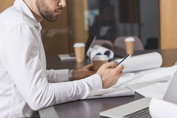 istock handsome architect with paper cup of coffee using smartphone at office 1009467750
