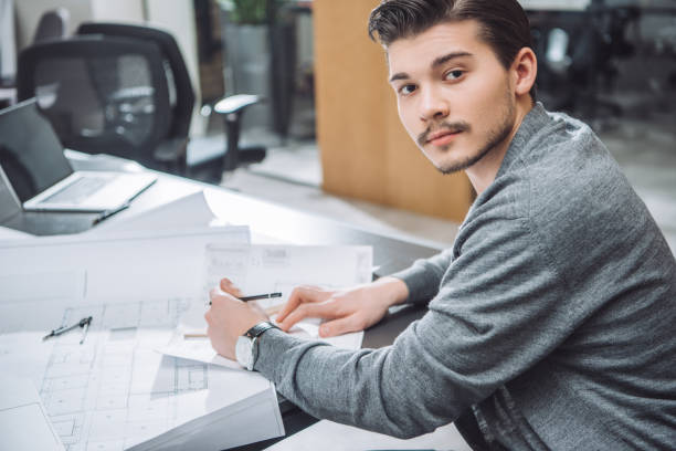 istock handsome young architect drawing building plans at office and looking at camera 1009474484