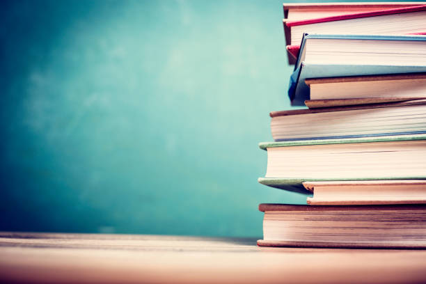 Textbooks on wooden school desk with chalkboard. Back to school.  Textbooks stacked on wooden school desk in front of green chalkboard.  Classroom setting. textbook stock pictures, royalty-free photos & images