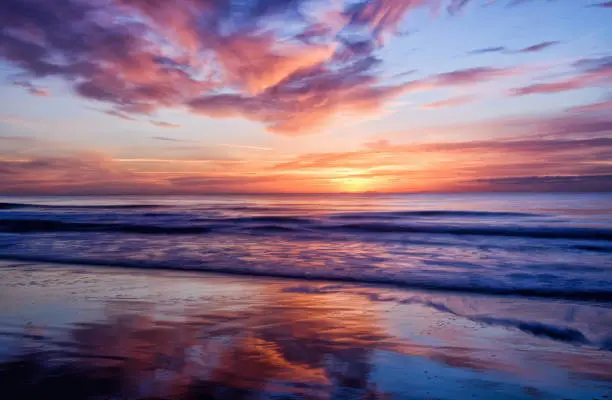 Long camera exposure captures the motion and reflections of the clouds in the wet sand and waves in the sea during a gorgeous sunrise in Myrtle Beach South Carolina.