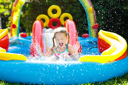 Child playing in inflatable baby pool. Kids swim, slide and splash in colorful garden play center. Happy little girl sliding and swimming with water toys on hot summer day. Family outdoor fun.
