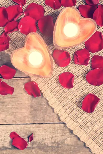 Enlightened candles in heart-shaped candleholders with red roses petals on rustic wooden background. St Valentines background. Romantic holiday concept. Top view. Vertical. Copy space at lower part. Warm creamy tone