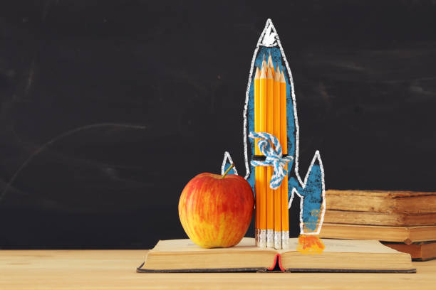 istock Back to school concept. rocket sketch and pencils over open book in front of classroom blackboard. 1009841430