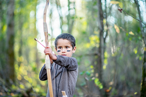 A young boy is outdoors in a forest. He is preparing to shoot a bow and arrow.