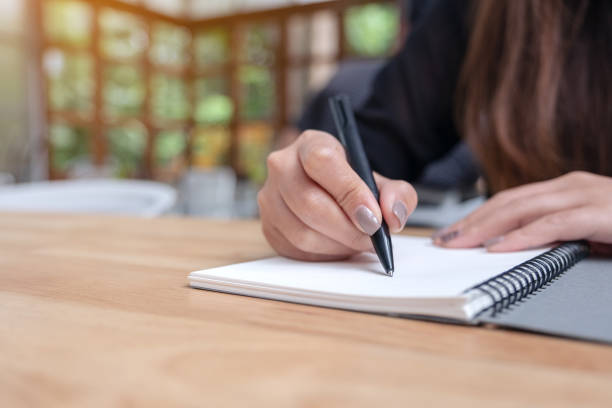 istock Closeup image of a woman's hand writing down on a white blank notebook on wooden table 1009942790