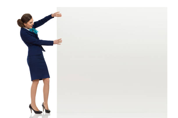 Young woman in blue formalwear and high heels is holding white big banner, looking at it and talking. Side view. Full length studio shot isolated on white.
