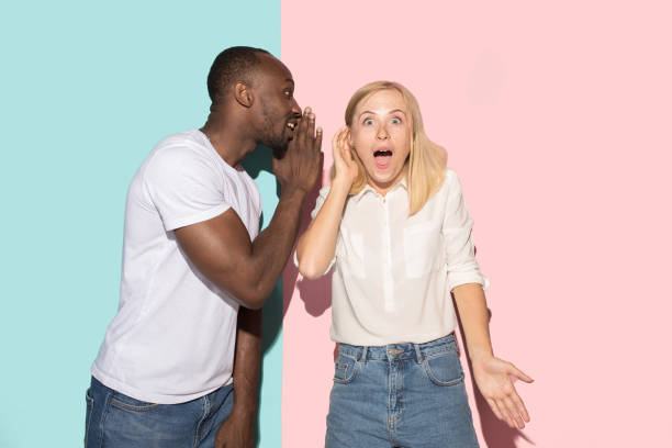 The young couple whispering a secret behind her hand over studio background Secret, gossip concept. Young couple whispering a secret behind hands. Business woman and afro man on trendy pink and blue studio background. Young emotional woman. Human emotions, facial expression concept. whisper men stock pictures, royalty-free photos & images