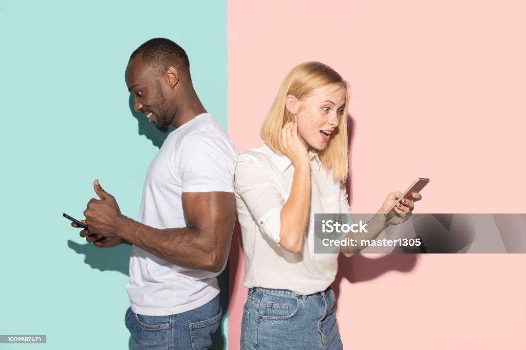 Mixed raced couple of students taking mobile phones. Caucasian girl and her African boyfriend posing at studio Mixed raced couple of students taking mobile phones. The surprised caucasian girl and her african boyfriend posing at studio . Communication and relationship concept Men Stock Photo
