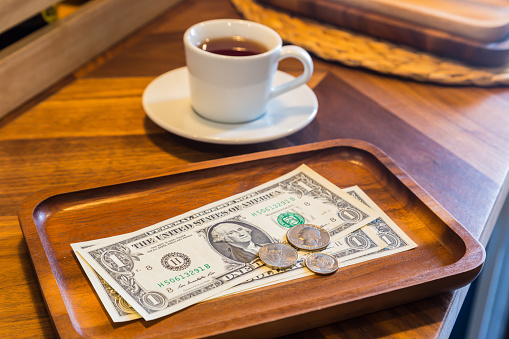 Dollar banknotes and US coins on wooden tray, on coffee shop table with a white cup of tea on background. Dim light room, modern and minimal style. Concepts for customer service, checking, money tips.
