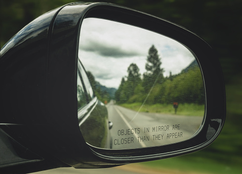Close-up of a rearview mirror with the text Objects in mirror are closer than they appear. Car on the road.