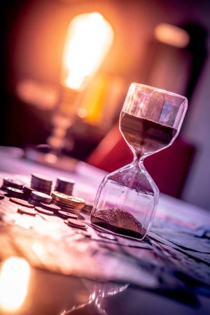 sand running through the shape of hourglass on table with banknotes and coins of international currency. time investment and retirement saving. urgency countdown timer for business deadline concept - hourglass time purple deadline imagens e fotografias de stock