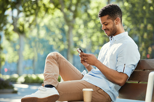 Happy man using smart phone while sitting on bench at sidewalk. Young male is relaxing in city on sunny day. Hipster is wearing casuals.