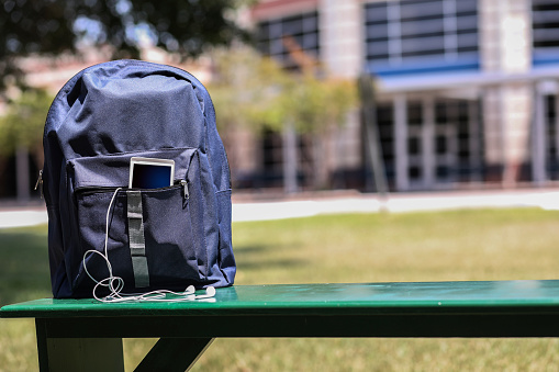 Various educational, learning objects on bench in front of a school building.  Items include:  backpack, cell phone, earbuds.