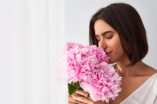 Portrait of young attractive dark-haired Caucasian woman enjoying scent of freshly picked pink peonies with her eyes closed