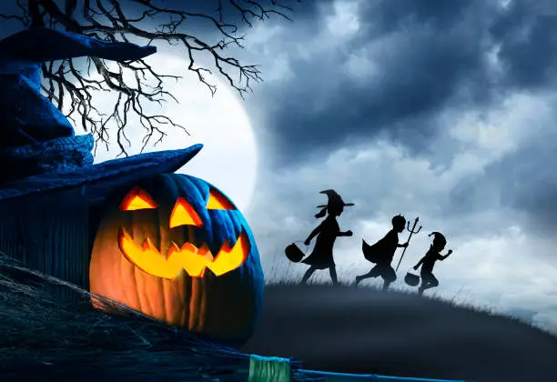 A jack o lantern, a witch's hat, and a broom stick, sit in front of three trick or treating children silhouetted against a cloudy moonlit sky on Halloween night.