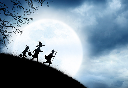 Three trick or treating children are silhouetted against a rising full moon as the make their way across grass covered hill.