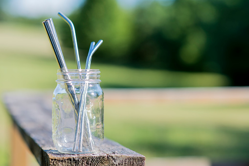 Reusable glass jar sitting on a wooden deck filled with stainless steel straws on a warm summer day outdoors.