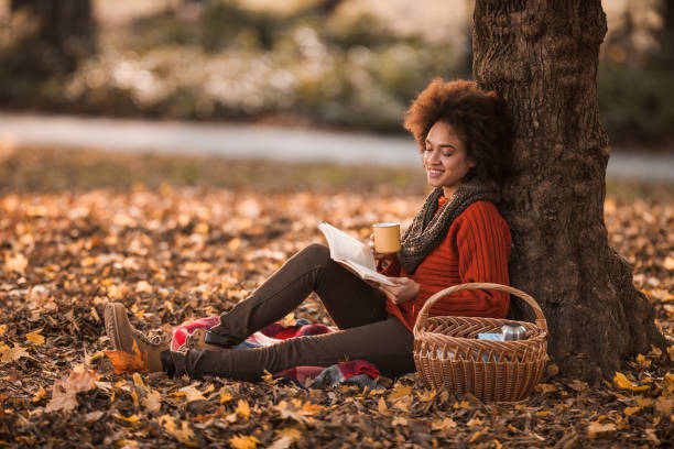 donna nera felice che legge un libro mentre si gode il picnic nella giornata autunnale. - enjoying a novel foto e immagini stock