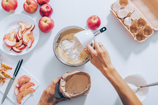 Top view woman adding flour into dough to prepare apple pie. Flat lay composition of kitchen table with ingredients for cooking seasonal fruit cake.