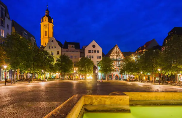 View over a fountain on the town square of Jena with historic buildings, outdoor restaurants and the city church (Stadtkirche) at night