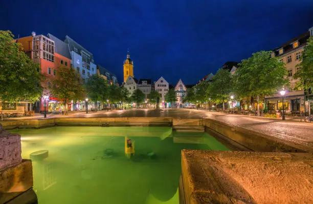 View over a fountain on the town square of Jena with historic buildings, outdoor restaurants and the city church (Stadtkirche) at night
