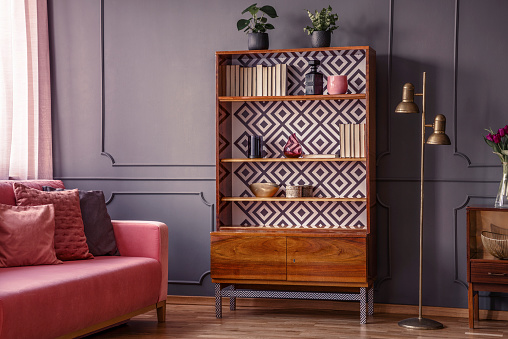 Renovated wooden bookcase with geometrical pattern and a brass floor lamp in a classy, dark living room interior with a comfortable pink settee