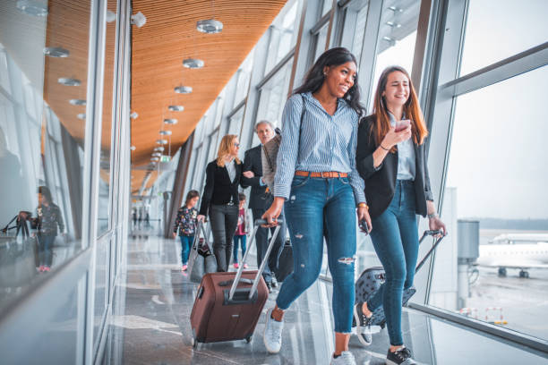 Female friends walking by window at airport Young woman showing mobile phone to friend while business people walking in background. Passengers are with luggage by window at airport. They are on tiled floor. passenger stock pictures, royalty-free photos & images