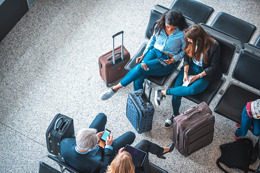 High angle view of passengers waiting for flights at departure area. People are sitting on seats at airport terminal. They are using technologies.