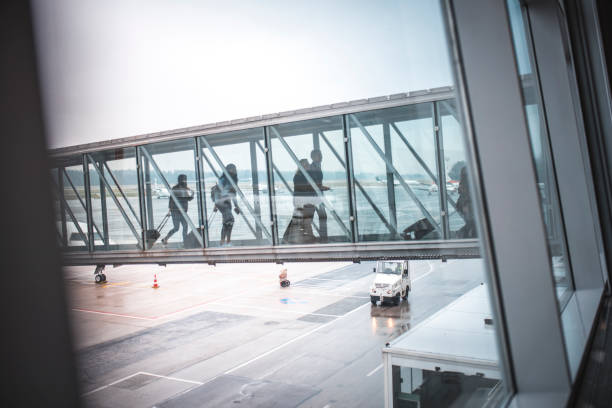 Passengers walking on footbridge at airport terminal. People with luggage are moving towards other part of building. They are seen through glass window.