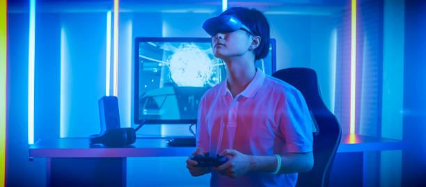istock East Asian Pro Gamer Wearing Virtual Reality Headset Plays Online Video Game Shooter using Joysticks / Controllers as Swords. Cool Retro Neon Colors in the Room. 1010650368