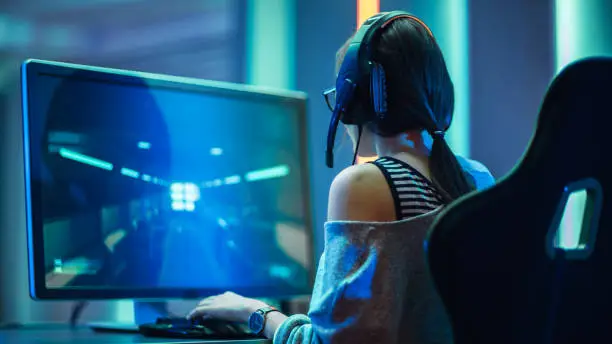 Photo of Shot of the Beautiful Pro Gamer Girl Playing in FPS Video Game on Her Personal Computer, Casual Cute Geek wearing Glasses and Headset. Neon Room. Online Cyber Games Internet Championship Event.