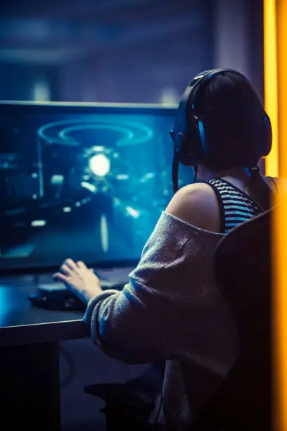 Back View Shot of the Beautiful Professional Gamer Girl Playing in Online First-Person Shooter Online Video Game on Her Personal Computer. Casual Cute Geek Girl in Dark Room Suddenly Lit by Neon Lights in Retro Arcade Style. Vertical Photo.