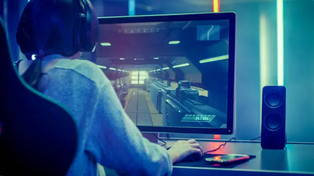 Back View Shot of the Beautiful Professional Gamer Girl Playing in Online First-Person Shooter Online Video Game on Her Personal Computer. Casual Cute Geek Girl Wearing Headset. Dark Room Suddenly Lit by Neon Lights in Retro Arcade Style.