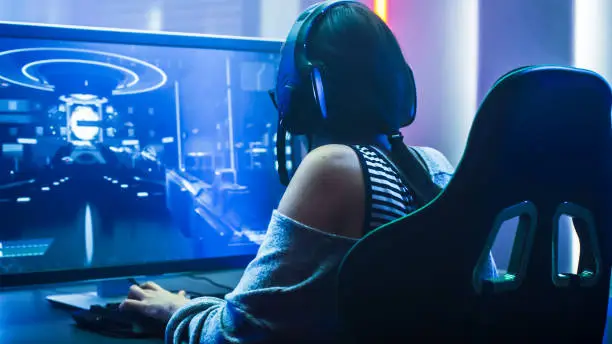 Shot of the Beautiful Pro Gamer Girl Playing in First-Person Shooter Online Video Game on Her Personal Computer. Casual Cute Geek wearing Glasses and Headset. Neon Room. eSport Cyber Games Internet Championship Event.