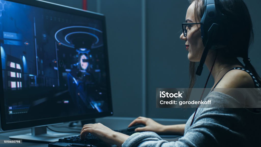 Shot of the Beautiful Professional Gamer Girl Playing in First-Person Shooter Online Video Game on Her Personal Computer. Casual Cute Geek wearing Glasses and Talking into Headset. In the Basement Gaming Club. Desktop PC Stock Photo