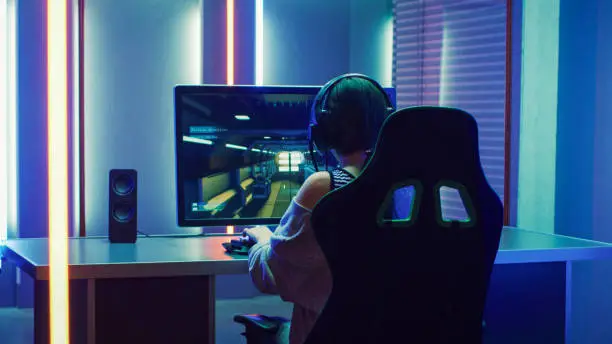 Beautiful Professional Gamer Girl Playing in First-Person Shooter Online Video Game on Her Personal Computer. Casual Cute Geek Girl Wearing Headset. Dark Room Suddenly Lit by Neon Lights in Retro Arcade Style.