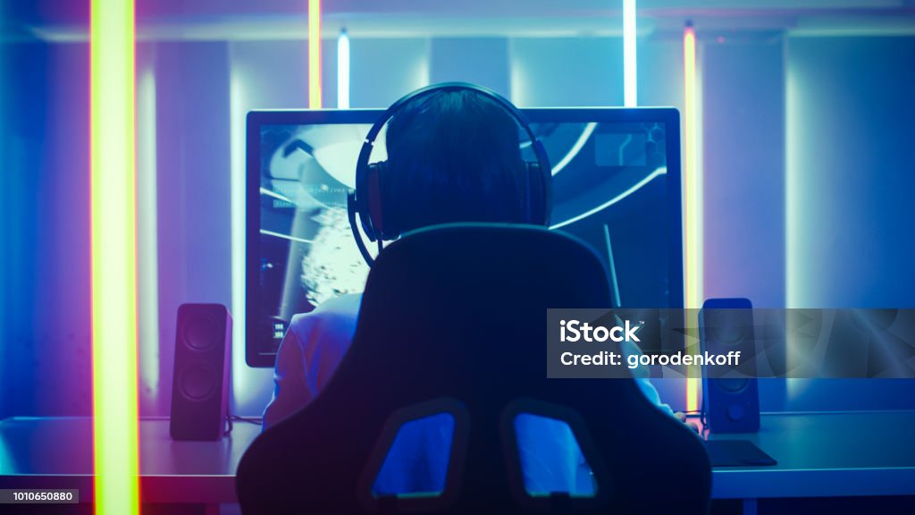 Back View Shot of the Professional Gamer Playing in First-Person Shooter Online Video Game on His Personal Computer. Room Lit by Neon Lights in Retro Arcade Style. Online Cyber e-Sport Internet Championship. eSports Stock Photo