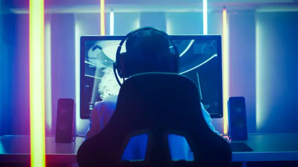 Back View Shot of the Professional Gamer Playing in First-Person Shooter Online Video Game on His Personal Computer. Room Lit by Neon Lights in Retro Arcade Style. Online Cyber e-Sport Internet Championship.